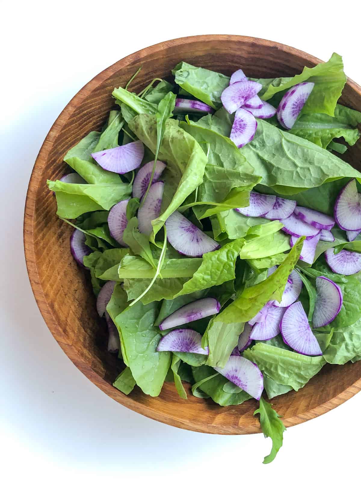 An image of a wooden bowl filled with fresh salad greens and purple daikon radishes.