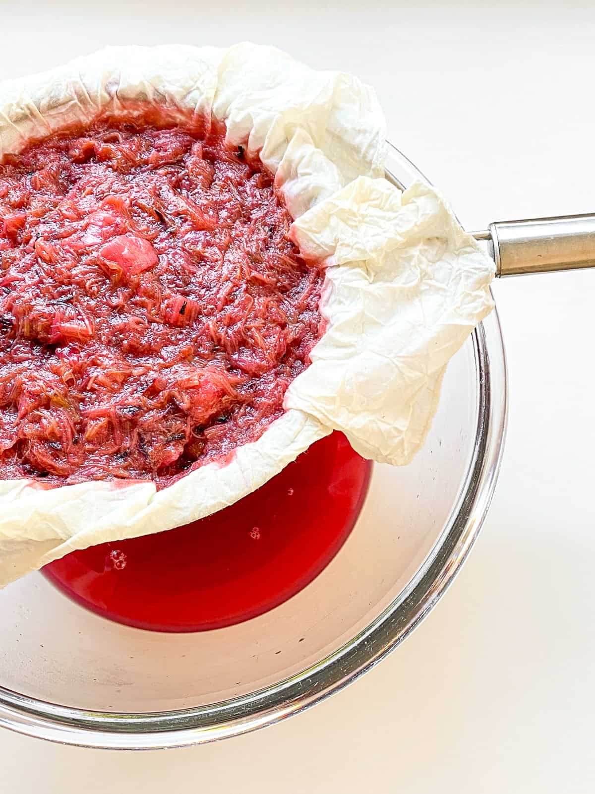 An image of cooked rhubarb being strained through cheesecloth and a mesh strainer over a glass bowl.
