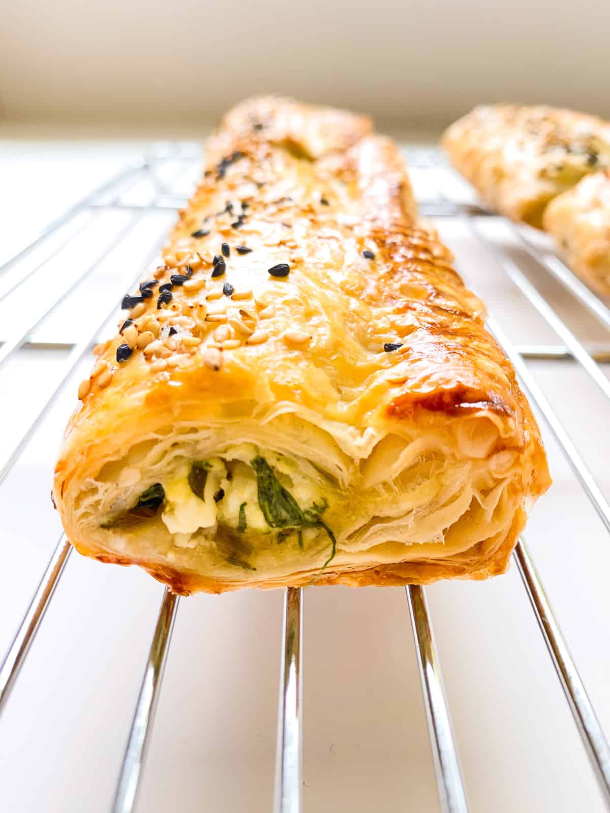 An image of a flaky greens filled puff pastry roll on a silver metal baking rack.