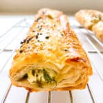 An image of a flaky greens filled puff pastry roll on a silver metal baking rack.