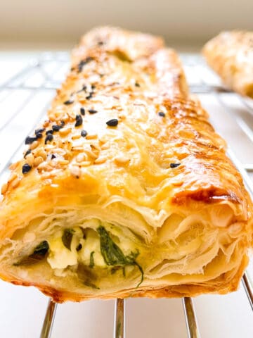 An image of a flaky greens filled puff pastry roll on a silver metal baking rack.