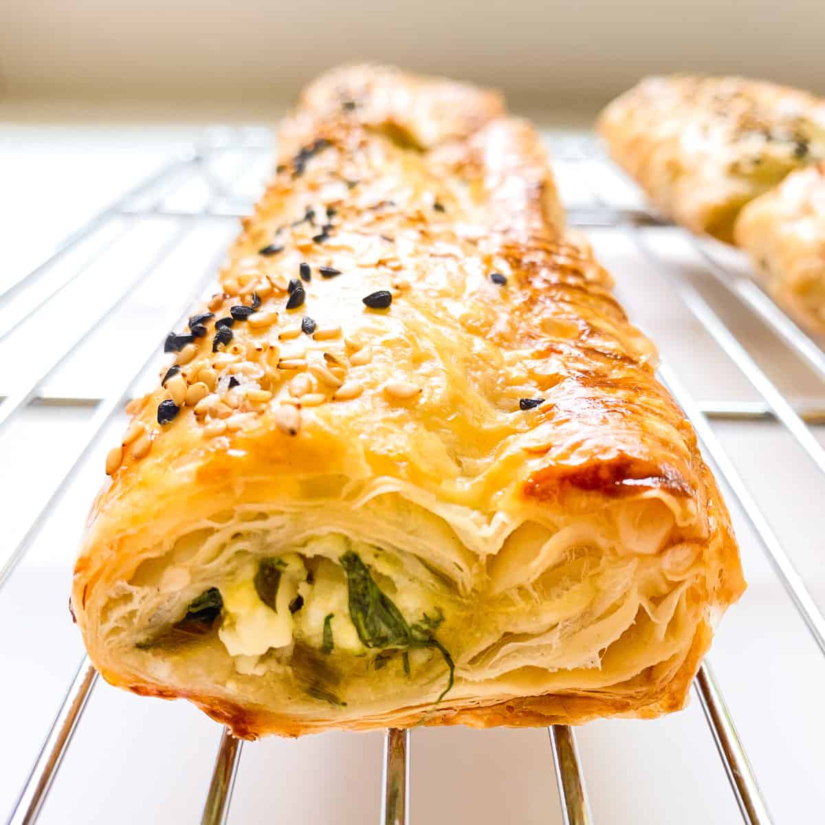 An image of a flaky greens filled puff pastry roll on a silver metal baking rack.