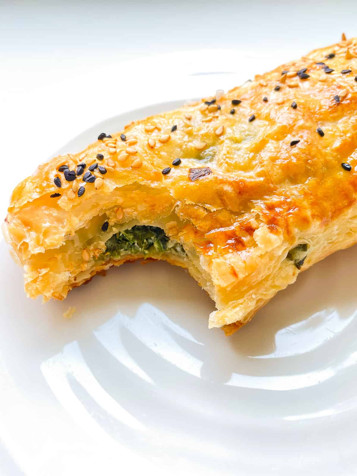 An image of a partially eaten flaky greens filled puff pastry roll on a white ceramic plate.