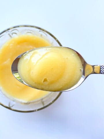 An image of a spoonful of applesauce near a glass bowl filled with applesauce.