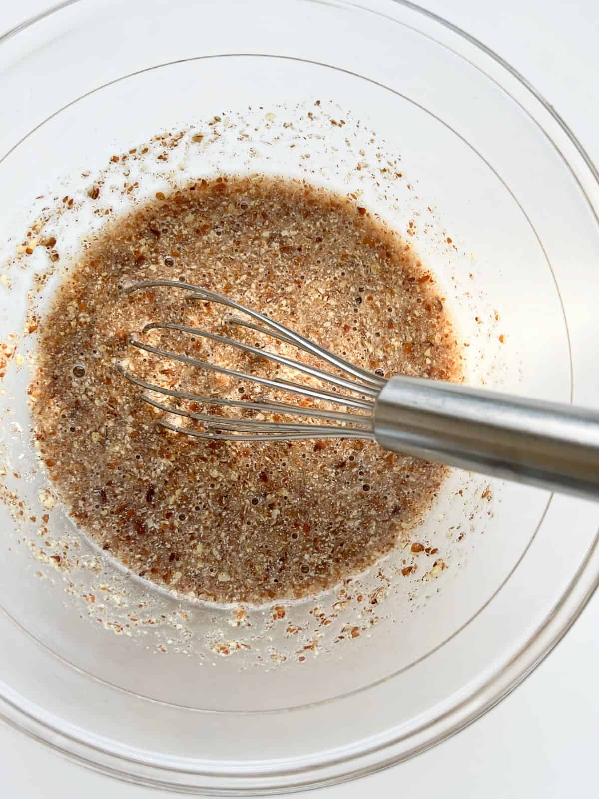 An image of ground flaxseed and water mixed together in a glass bowl with a silver whisk.