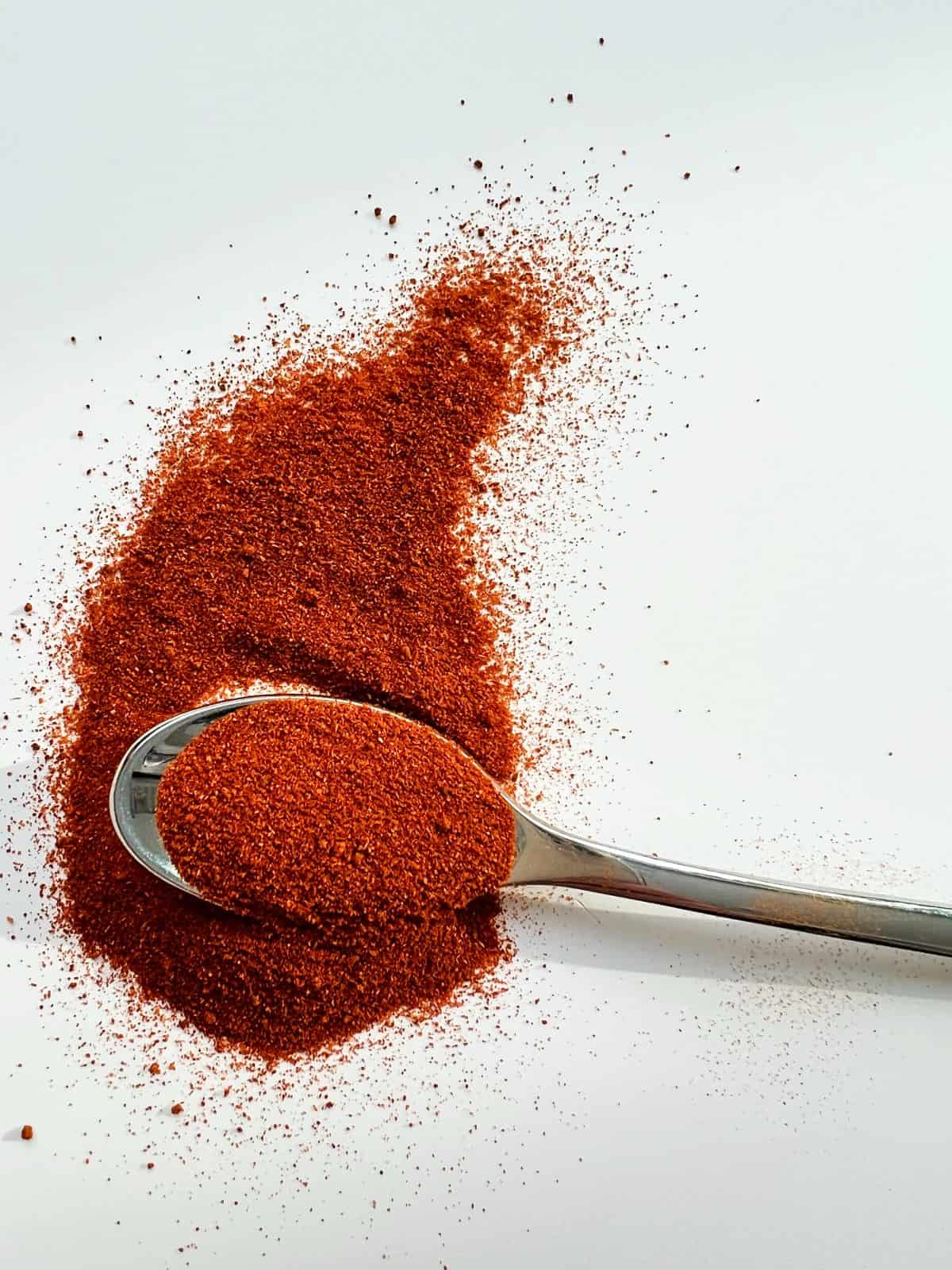 An image of spanish smoked paprika on a silver spoon and scattered against a white countertop.