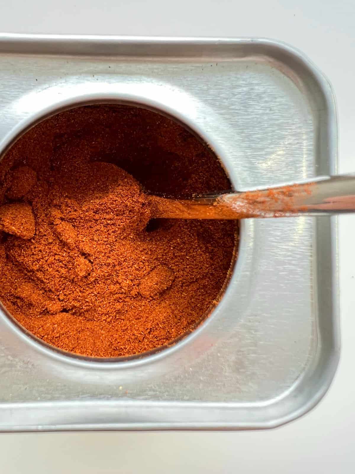 An image of spanish smoked paprika in a small tin, with a silver spoon in the red powder.
