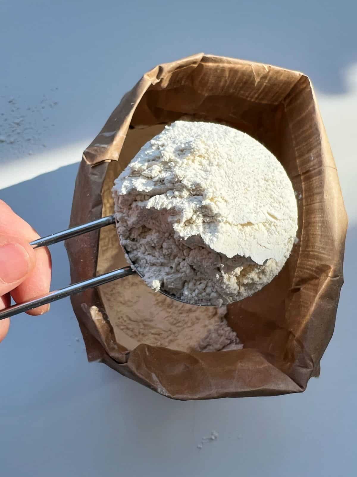 An image of a hand holding a cup of whole wheat pastry flour, with the brown paper flour bag visible in the background.