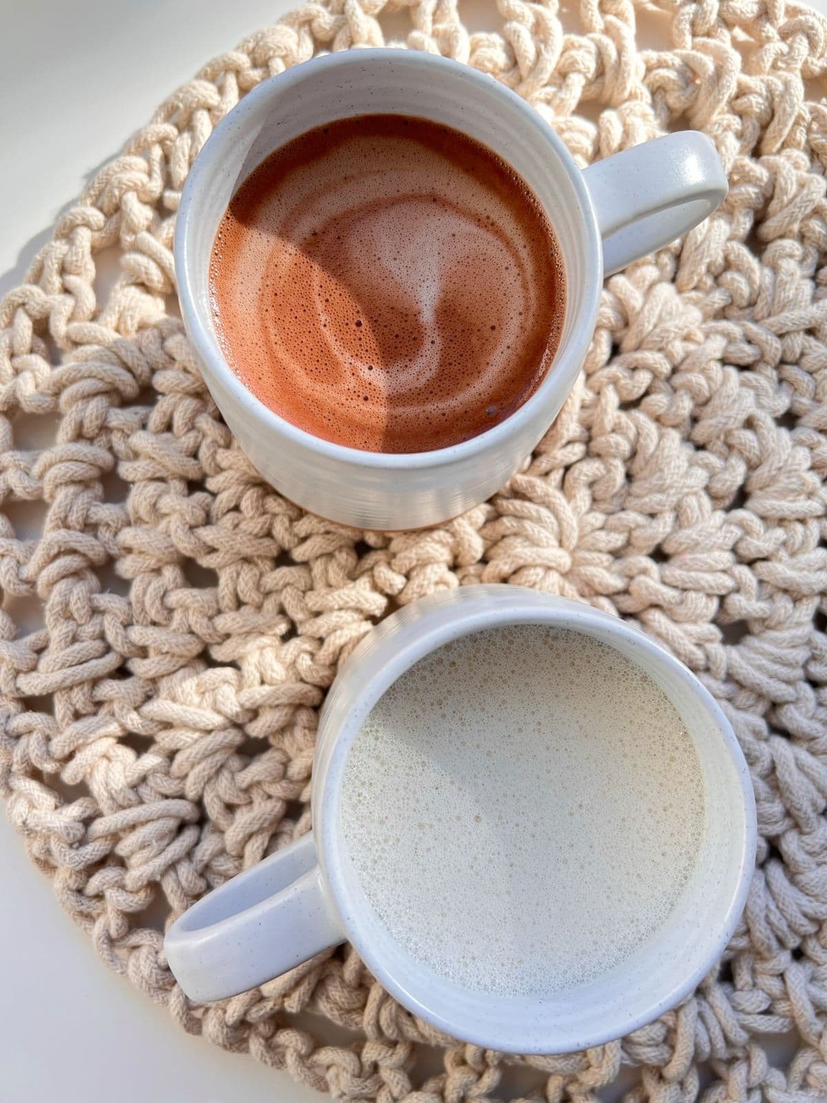 Two ceramic mugs of hot malted oat drinks, sitting on top of an oatmeal coloured placemat, one mug contains Hot Malted Cacao, and one mug contains Hot Malted Oat Milk.