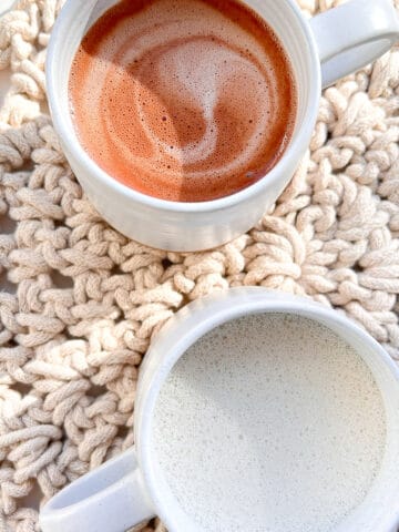 Two ceramic mugs of hot malted oat drinks, sitting on top of an oatmeal coloured placemat, one mug contains Hot Malted Cacao, and one mug contains Hot Malted Oat Milk.