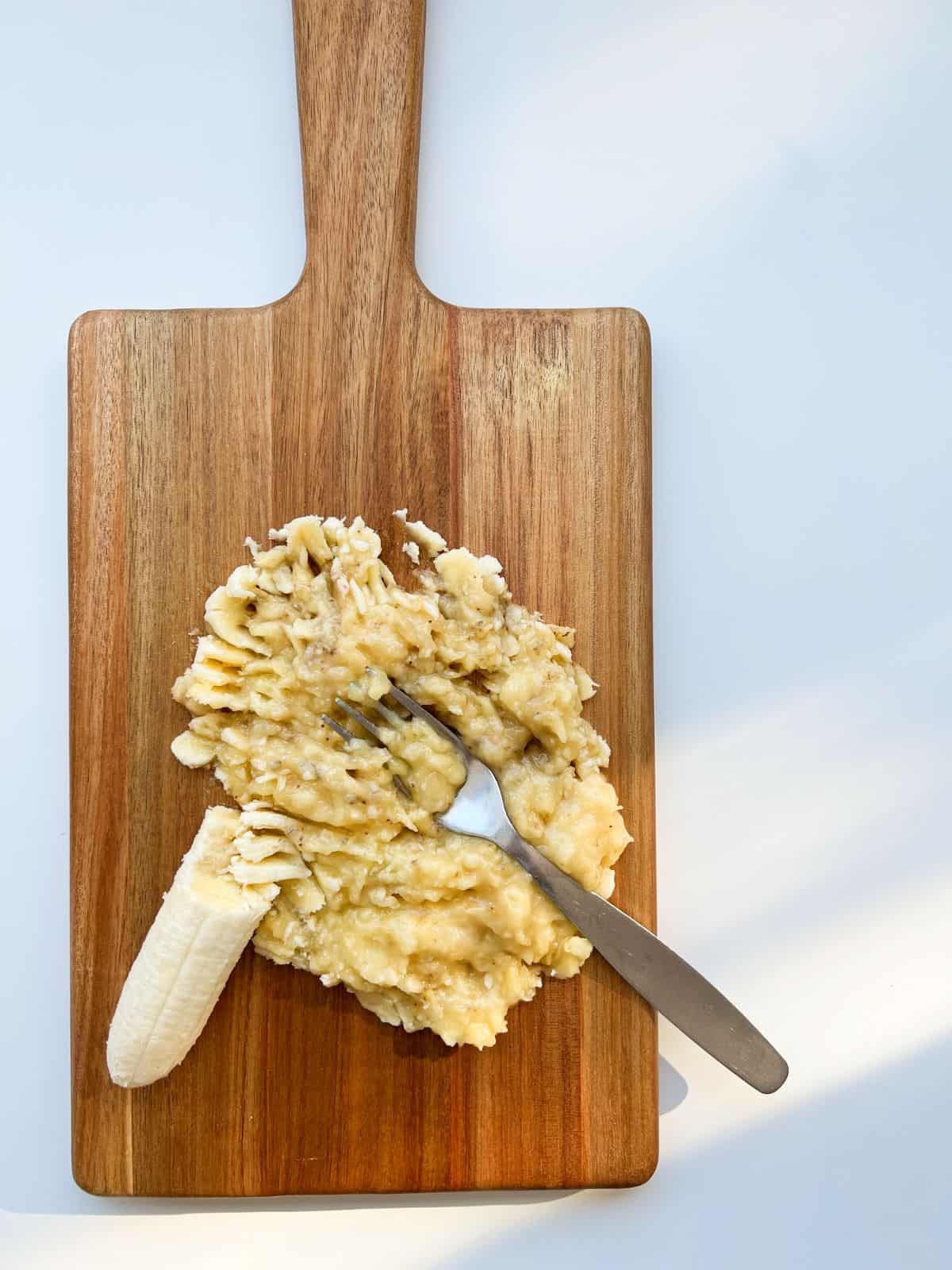 An image of banana mashed with a fork on a wooden cutting board.