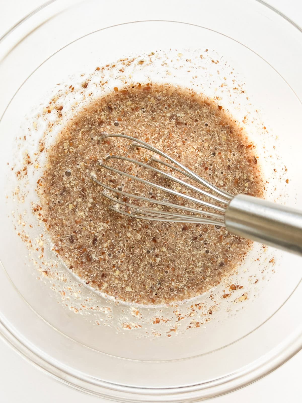 An image of ground flaxseed and water mixed together in a glass bowl with a silver whisk.