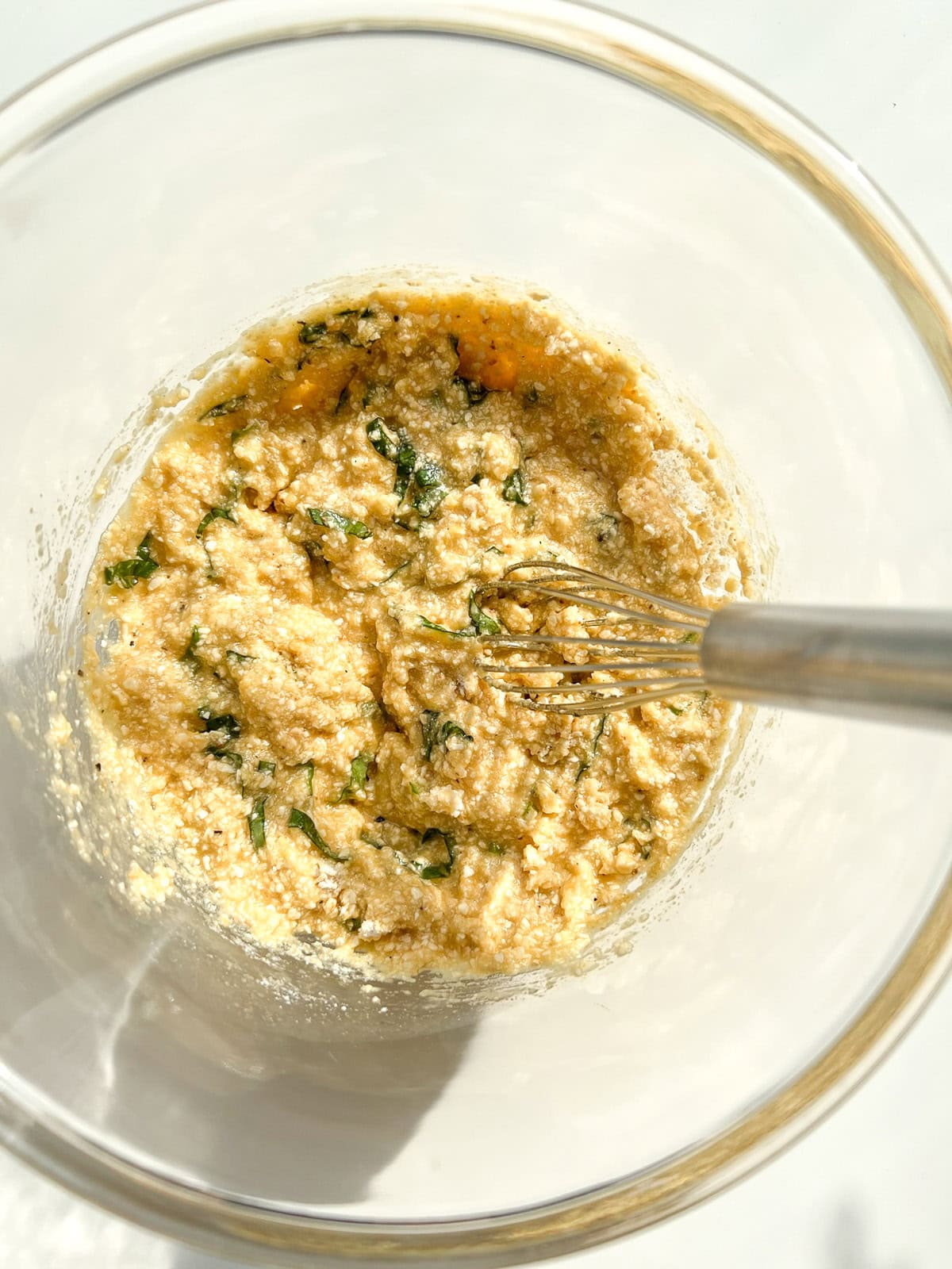 The matzo ball mixture being mixed with a small stainless steel whisk inside a glass bowl.