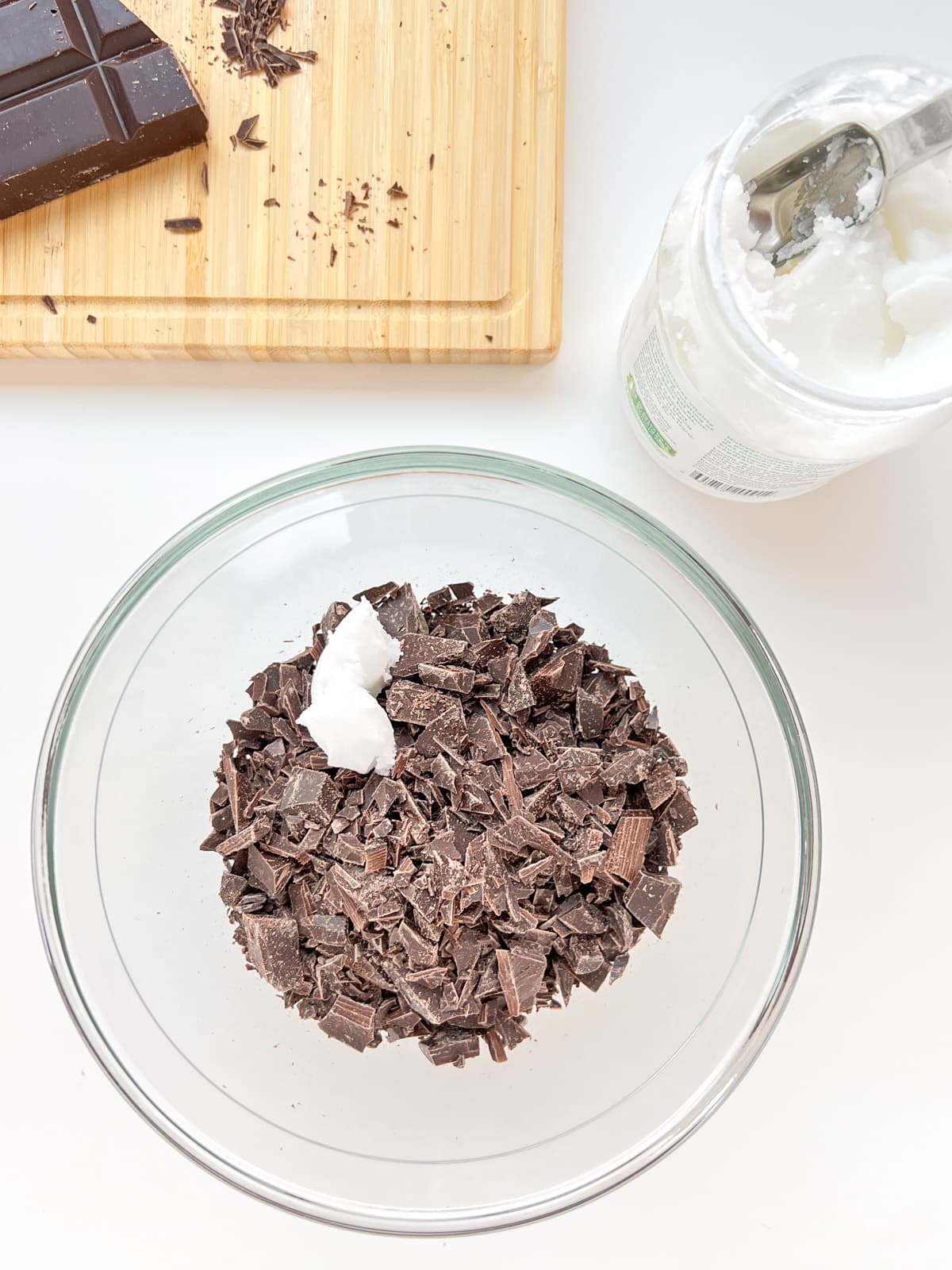 An image of a glass bowl containing chopped chocolate and coconut oil, with chocolate and coconut oil visible in the background.