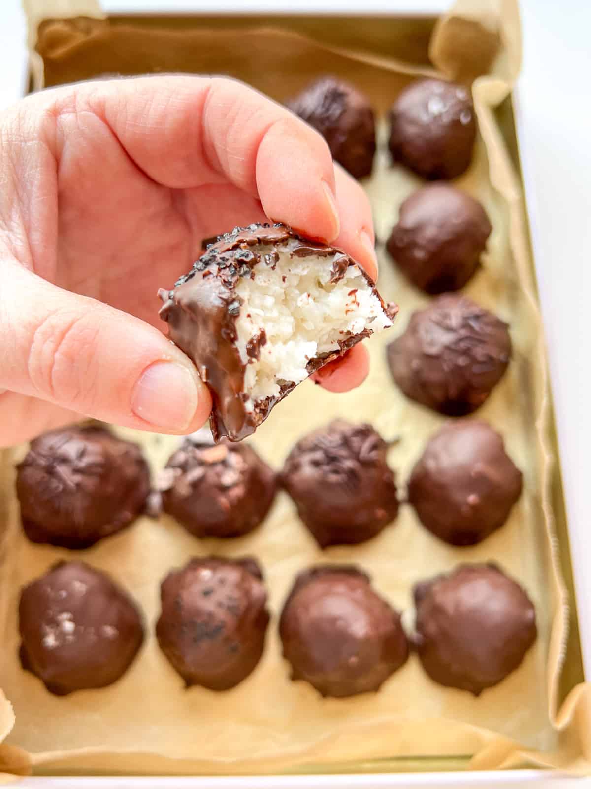 An image showing a hand holding a half eaten Chocolate Coconut Bonbon with the tin filled with more bonbons in the background.