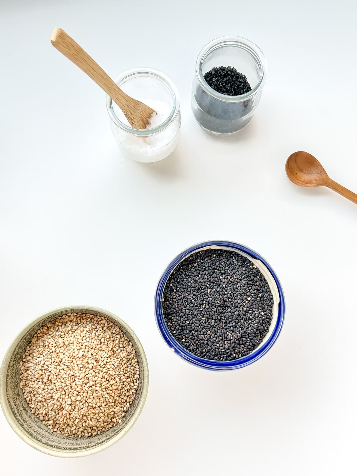 An image of four small containers against a white counter, containing white sesame seeds, black sesame seeds, sea salt, and black salt.