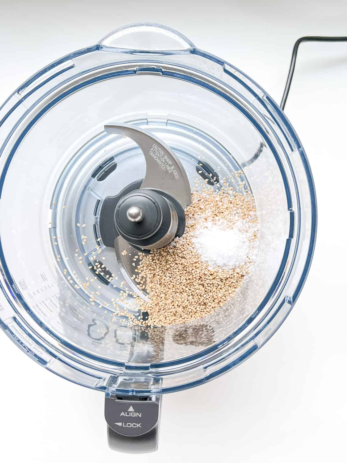 A close up of the bowl of a food processor containing sesame seeds and salt.