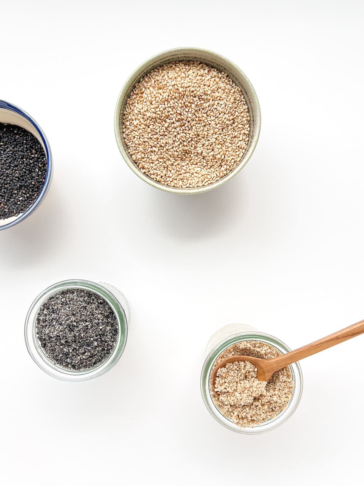 An image of four small containers against a white counter, containing white sesame seeds, black sesame seeds, black sesame seed salt, and white sesame seed salt.