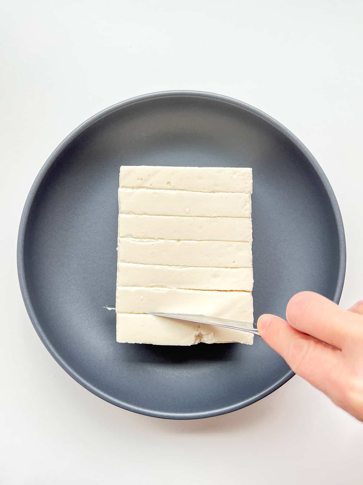 An image of tofu in a grey dish, with a hand shown cutting the tofu into slices.