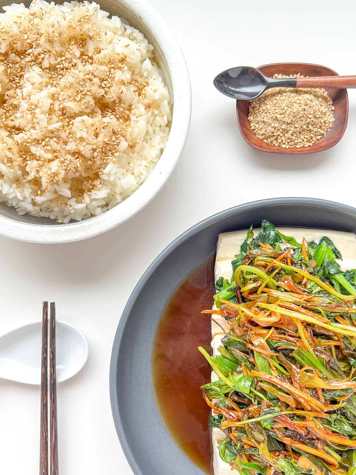 An image showing a meal laid out on a white counter, consisting of a dish of Steamed Silken Tofu with Ginger and Fragrant Greens, Steamed Rice sprinkled with Roasted Sesame Seed Salt, a spoon, chopsticks and a dish of Sesame Seed Salt.