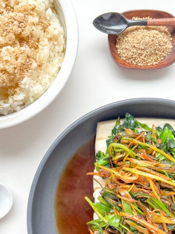 An image showing a meal laid out on a white counter, consisting of a dish of Steamed Silken Tofu with Ginger and Fragrant Greens, Steamed Rice sprinkled with Roasted Sesame Seed Salt, a spoon, chopsticks and a dish of Sesame Seed Salt.