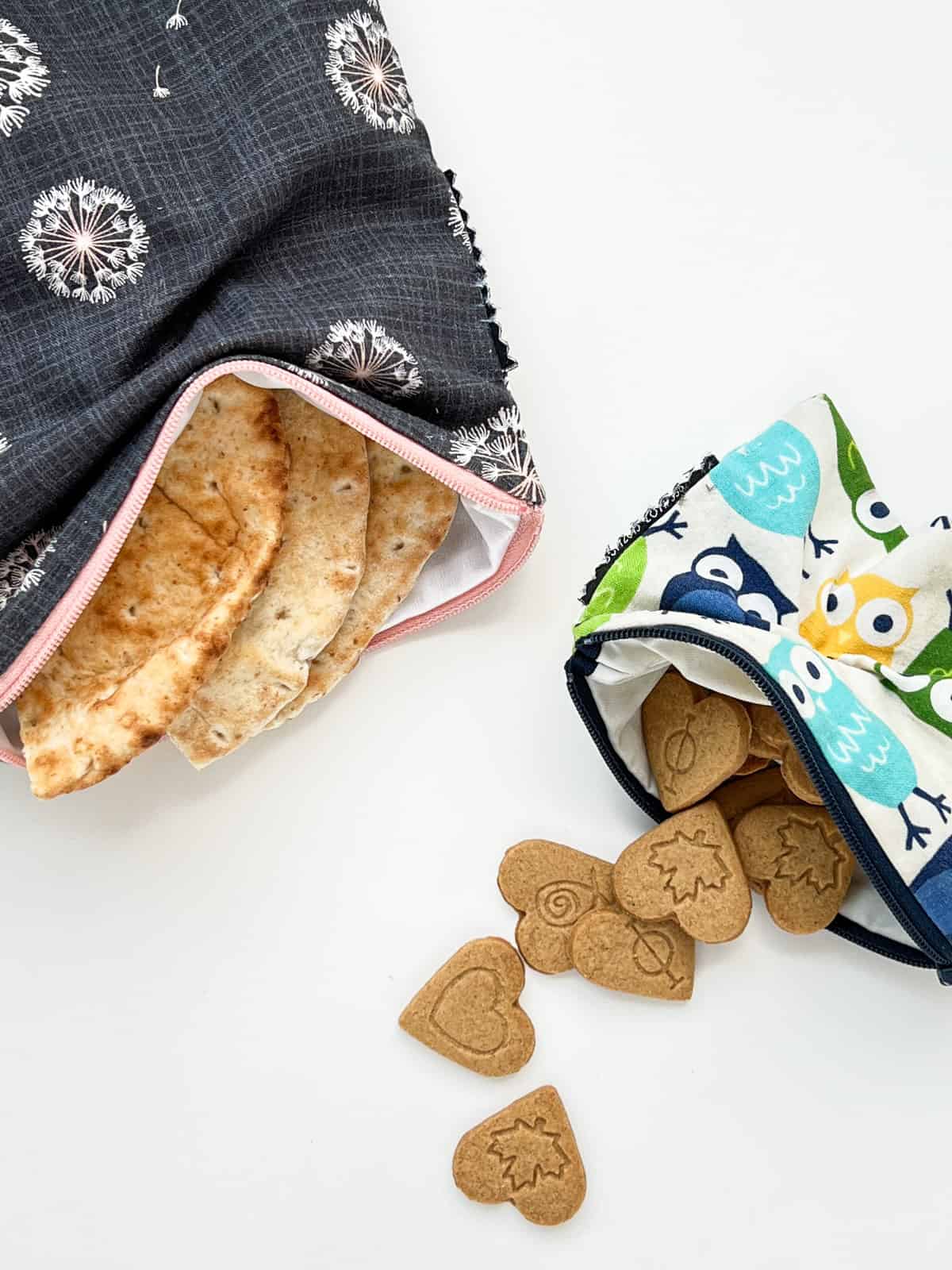 An image of two sizes of reusable fabric pouches that are used for food and snacks; on contains pita bread and the other contains small heart shaped cookies.