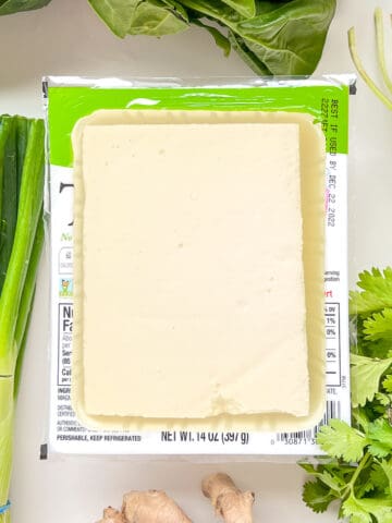 An image of silken tofu in an open container, surrounded by green vegetables and ginger.