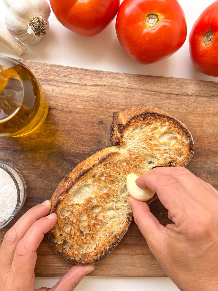 An image of a woman's hand rubbing a garlic clove on top of a toasted slice of bread.