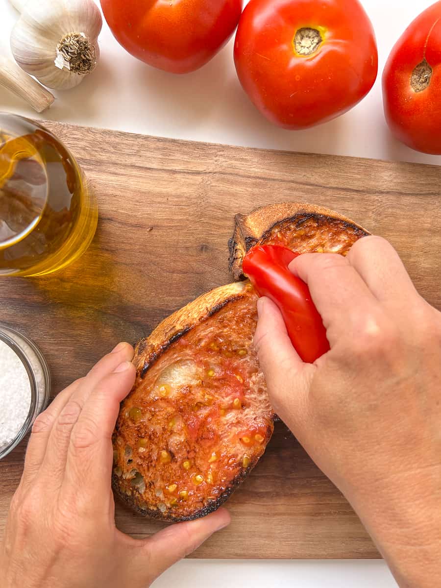An image of a woman's hand rubbing a cut half of tomato on top of a slice of bread.