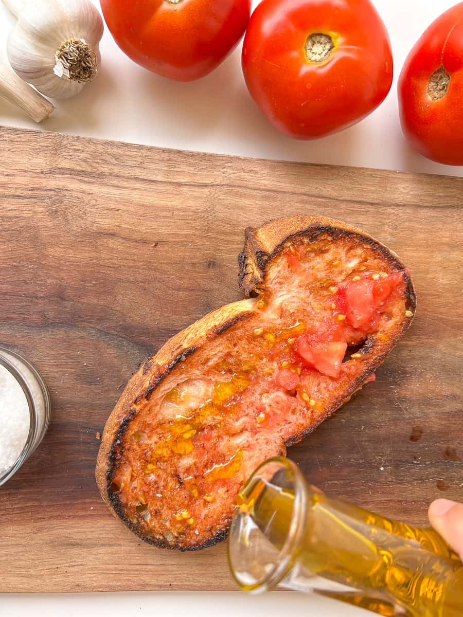 An image of a woman's hand pouring olive oil from a glass container onto the surface of a piece of Pan con Tomate.