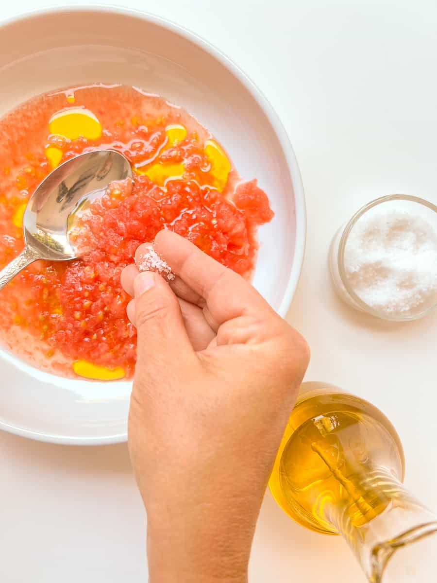 An image of a woman's hand sprinkling salt into a dish of tomato topping for Pan con Tomate.