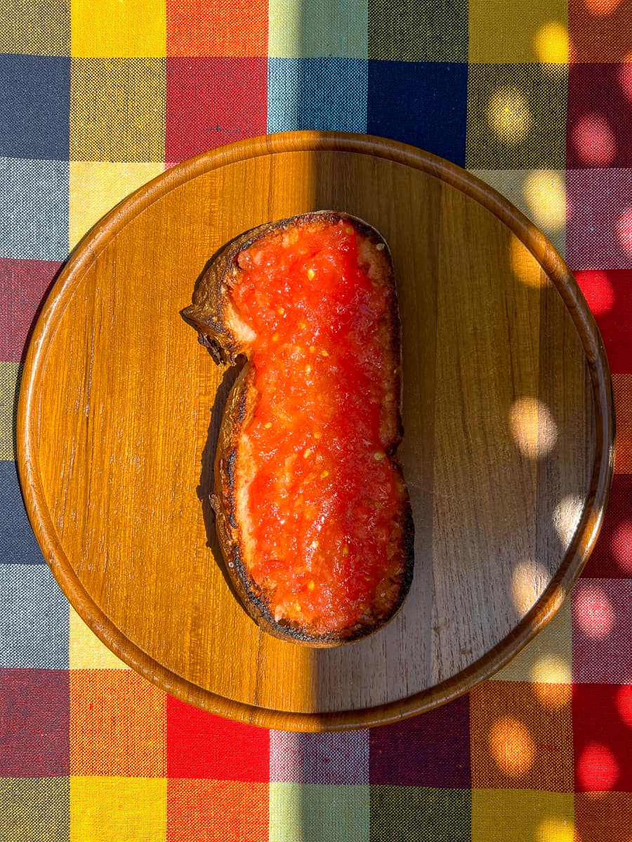An image of a piece of Pan con Tomate, on a wooden dish, on top of a chequered tablecloth.