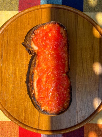 An image of a piece of Pan con Tomate, on a wooden dish, on top of a chequered tablecloth.