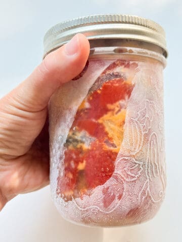 An image of a woman's hand holding a mason jar that has been frozen; frozen roasted tomatoes are visible inside.