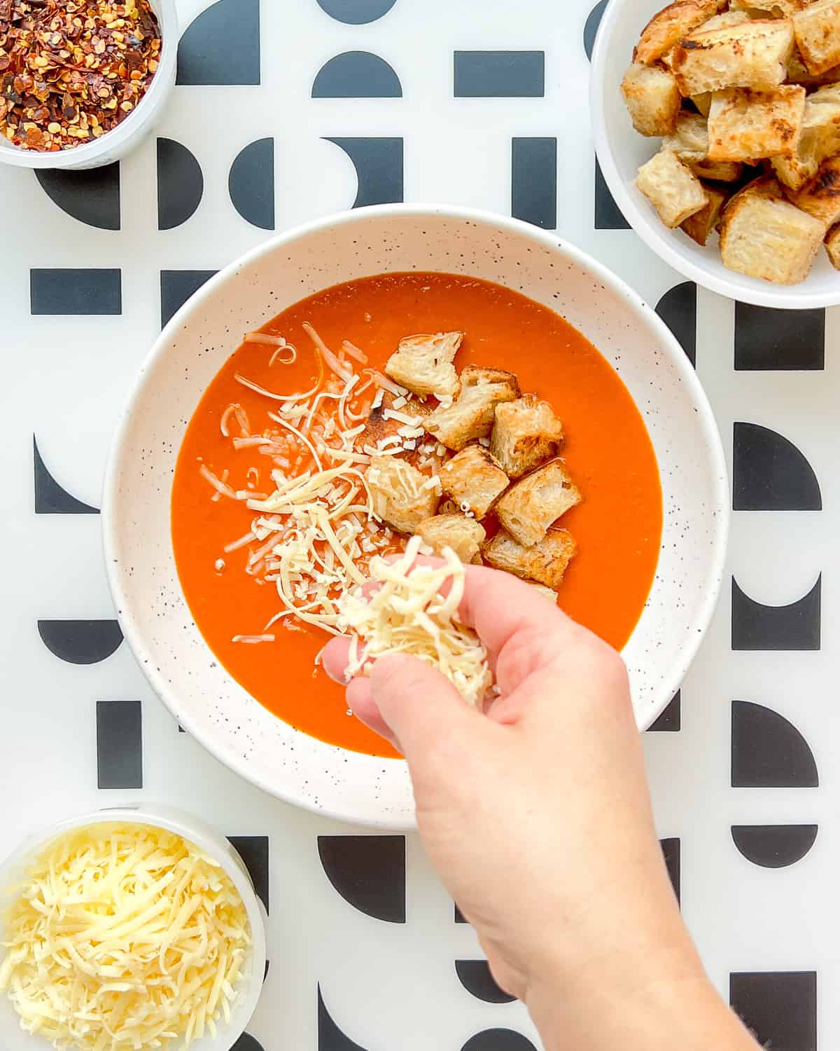 An image of roasted tomato soup being garnished with shredded cheese.