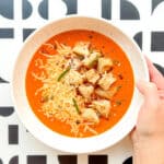 An image of Roasted Tomato and Garlic Soup with Pan-Crisped Croutons ready to eat with a graphic black and white background, with a woman's hand holding the soup bowl..