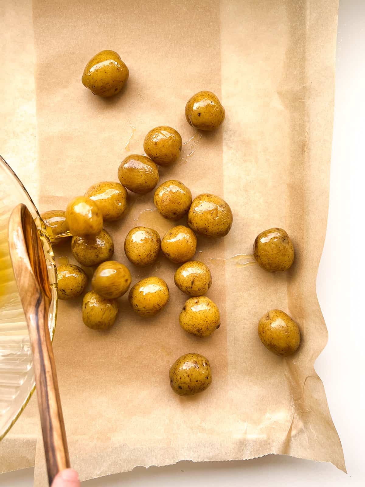 An image of oil coated potatoes being scooped onto a parchment lined roasting pan.