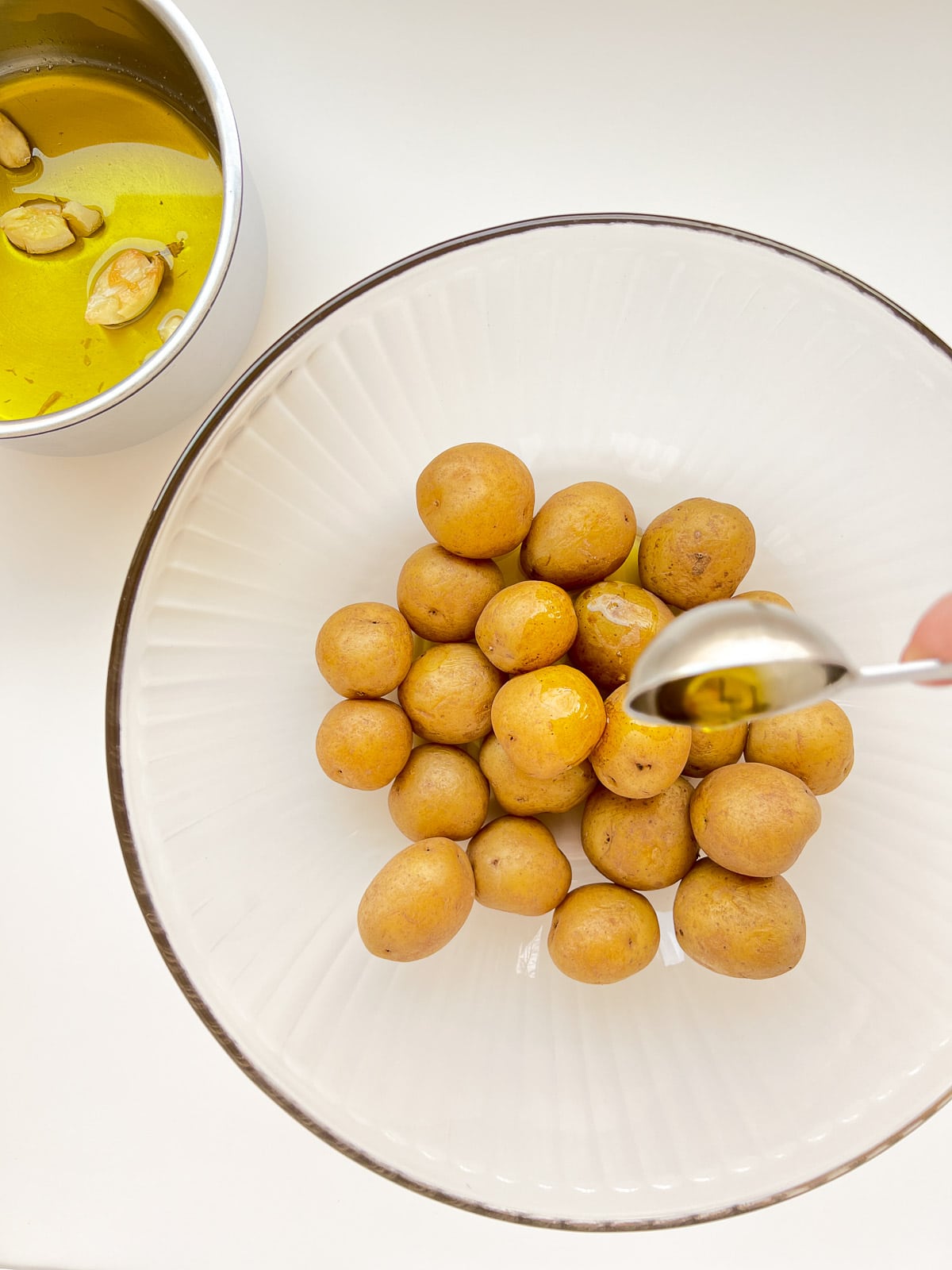 An image of garlic oil being spooned onto small potatoes in a glass bowl.