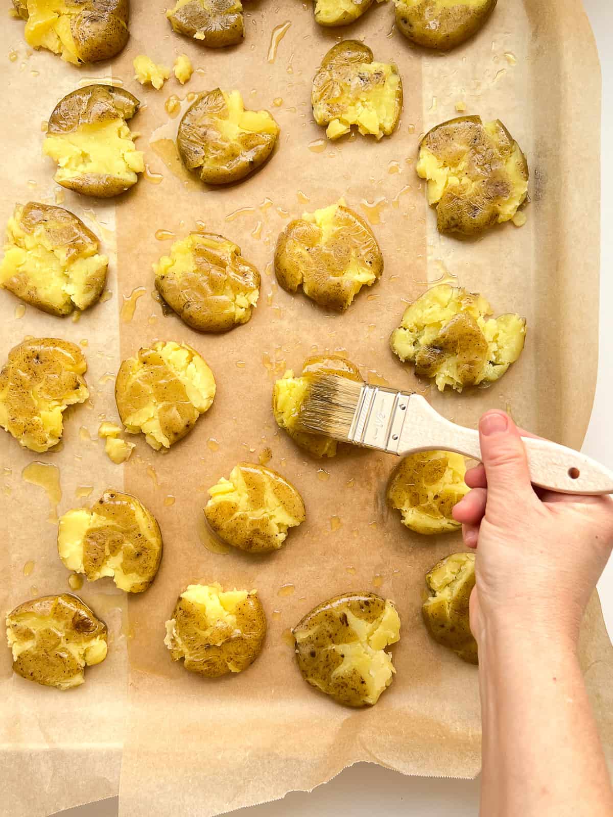 An image of potatoes being brushed with garlic oil after they have been smashed onto a roasting pan.