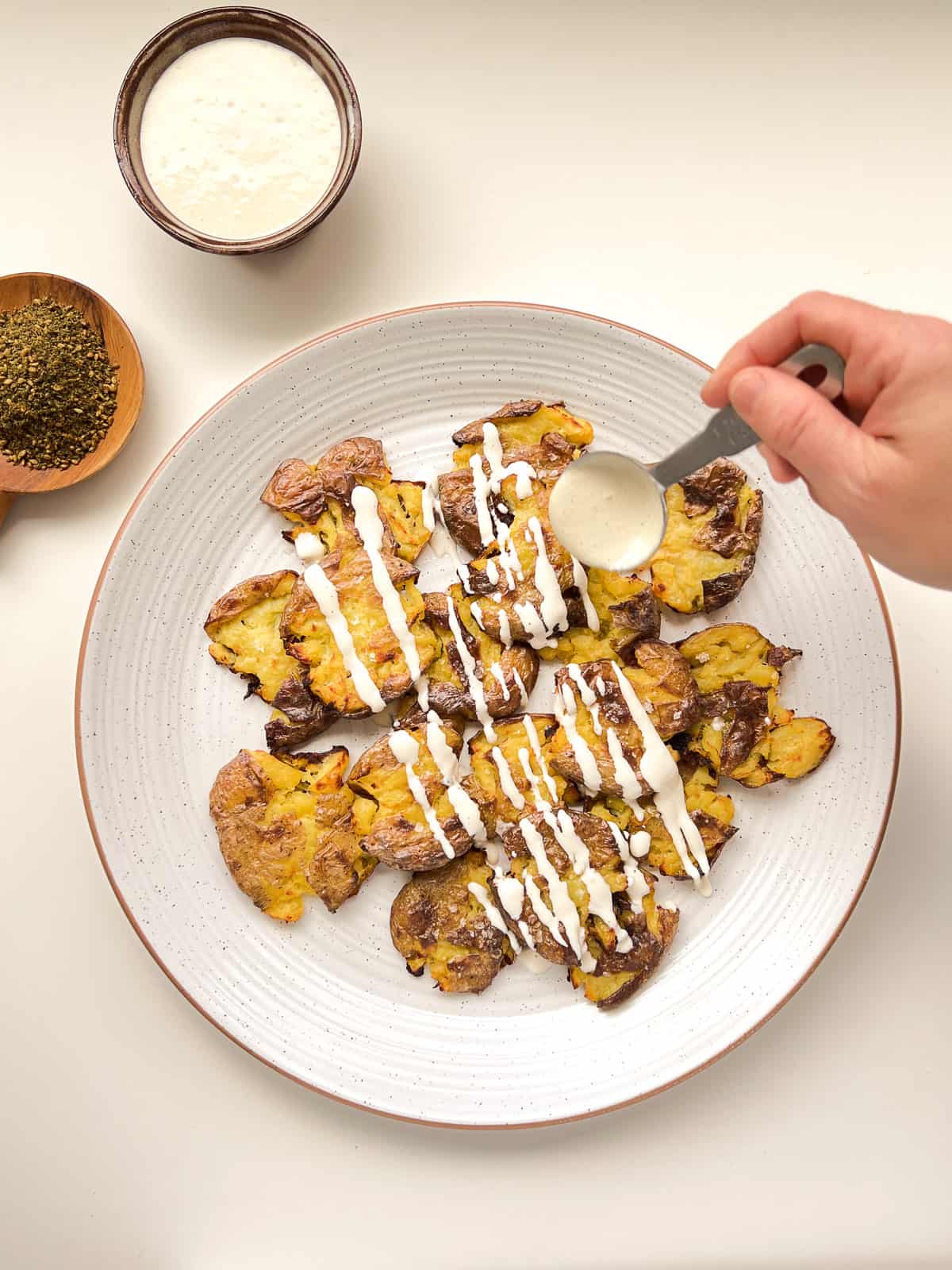 An image of a hand drizzling tahini over crispy smashed potatoes.