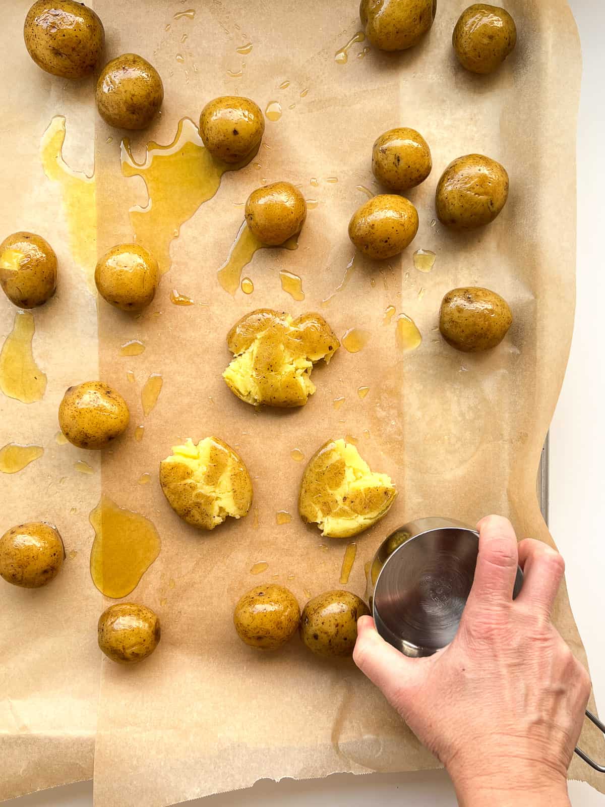 An image of a hand smooshing small boiled potatoes using the back of a metal measuring cup.