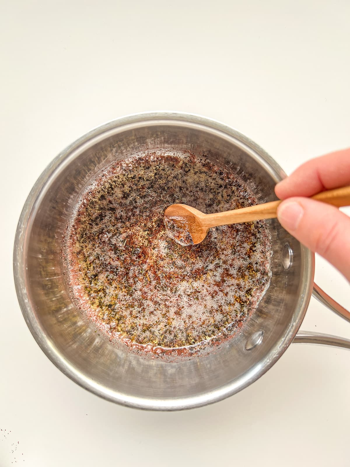 An image of a hand stirring a small wooden spoon that sits in a small stainless steel pot filled with herbed butter.