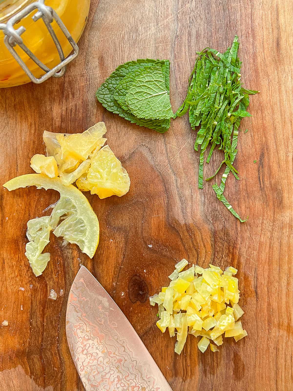 An image of preserved lemon being cut, and mint being slivered, on a wooden cutting board.
