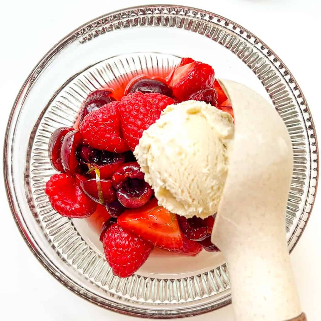 An image of Summer Fruit Salad in a glass bowl with a scoop of vanilla ice cream being scooped on top.