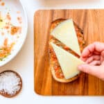 An image of a woman's hand placing a piece of manchego cheese on top of a piece of Pan con Tomate with Fresh Herbs and Manchego.