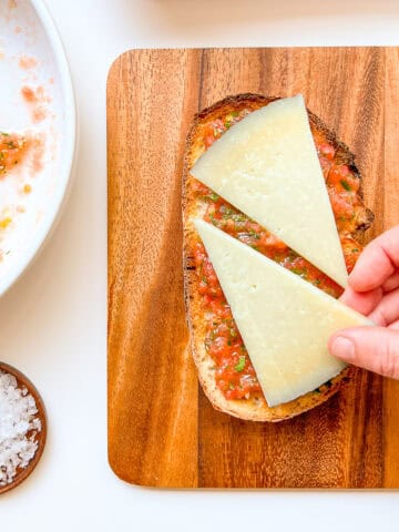 An image of a woman's hand placing a piece of manchego cheese on top of a piece of Pan con Tomate with Fresh Herbs and Manchego.